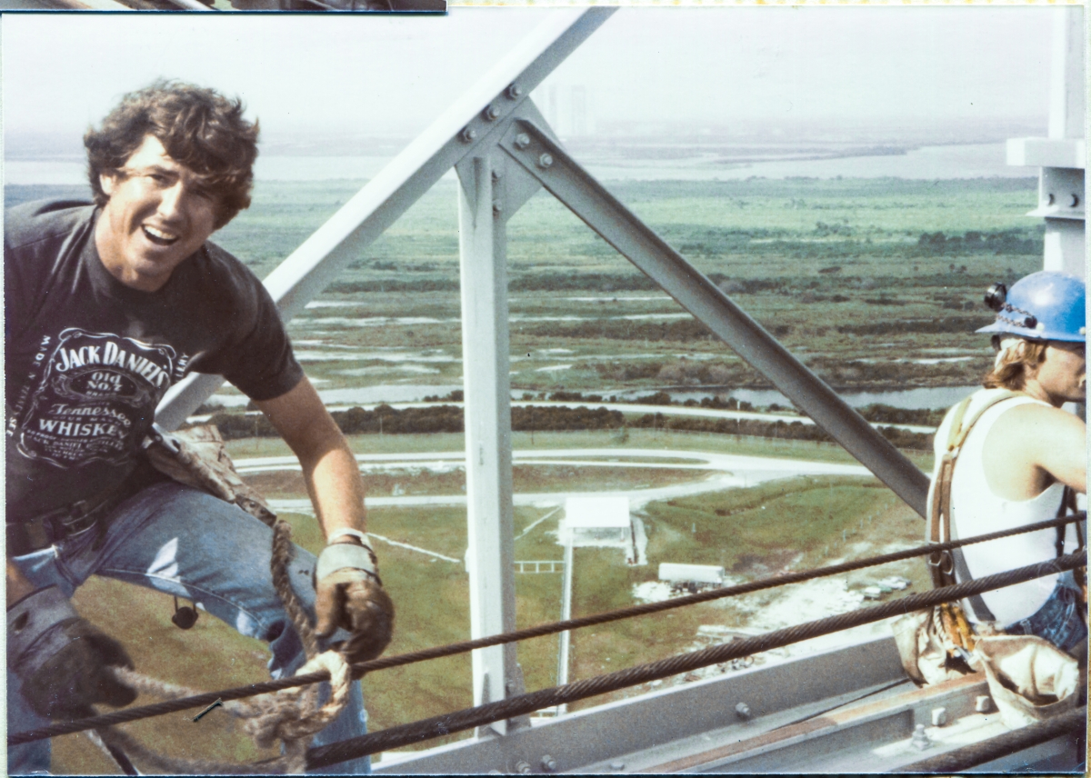 Image 062. Union Ironworker Steve Parker, from Local 808, working for Ivey Steel at Space Shuttle Launch Complex 39-B, Kennedy Space Center, Florida, turns and laughs incredulously at the photographer an instant after the strong wind which was blowing this day snatched his hardhat from his head and sent it to the bottom of the Flame Trench, 300 feet below, while he was engaged in the installation of the TPS Inspection Spider Basket Trolley Support Extensible Pipe Boom to the Hammerhead Crane. Photo by James MacLaren.